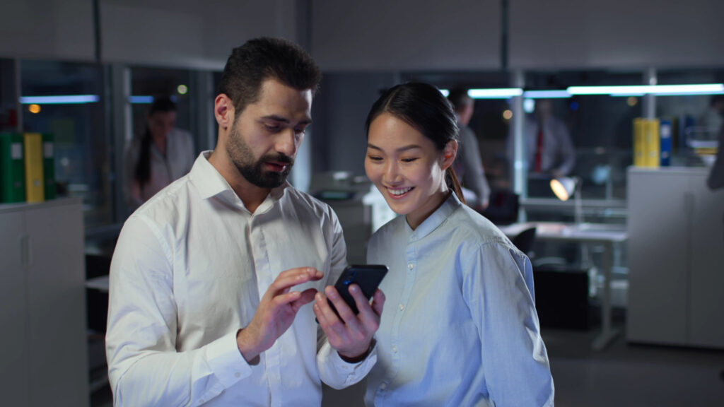Diverse colleagues using smartphone and talking standing in office. Multiracial business people work at night in modern office.